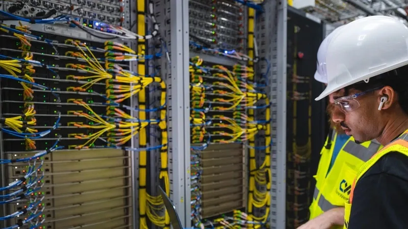 Two CoreWeave technicians in hard hats and safety vests reviewing a laptop before a server rack of yellow fiber cabling