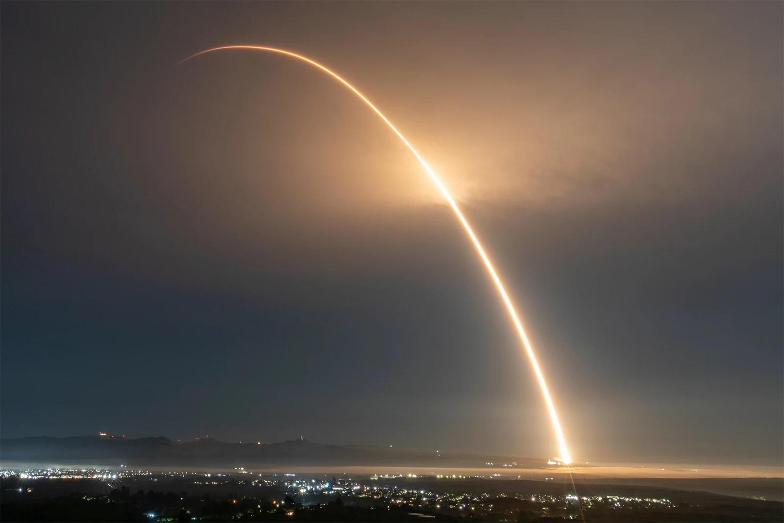 Long-exposure photo shows a Falcon 9 commercial space launch leaving a curved bright trail over a coastline