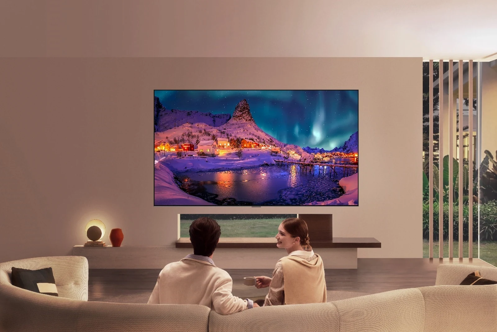 LG OLED evo TV on a living room wall shows an aurora over a snowy village as two people watch from a sofa