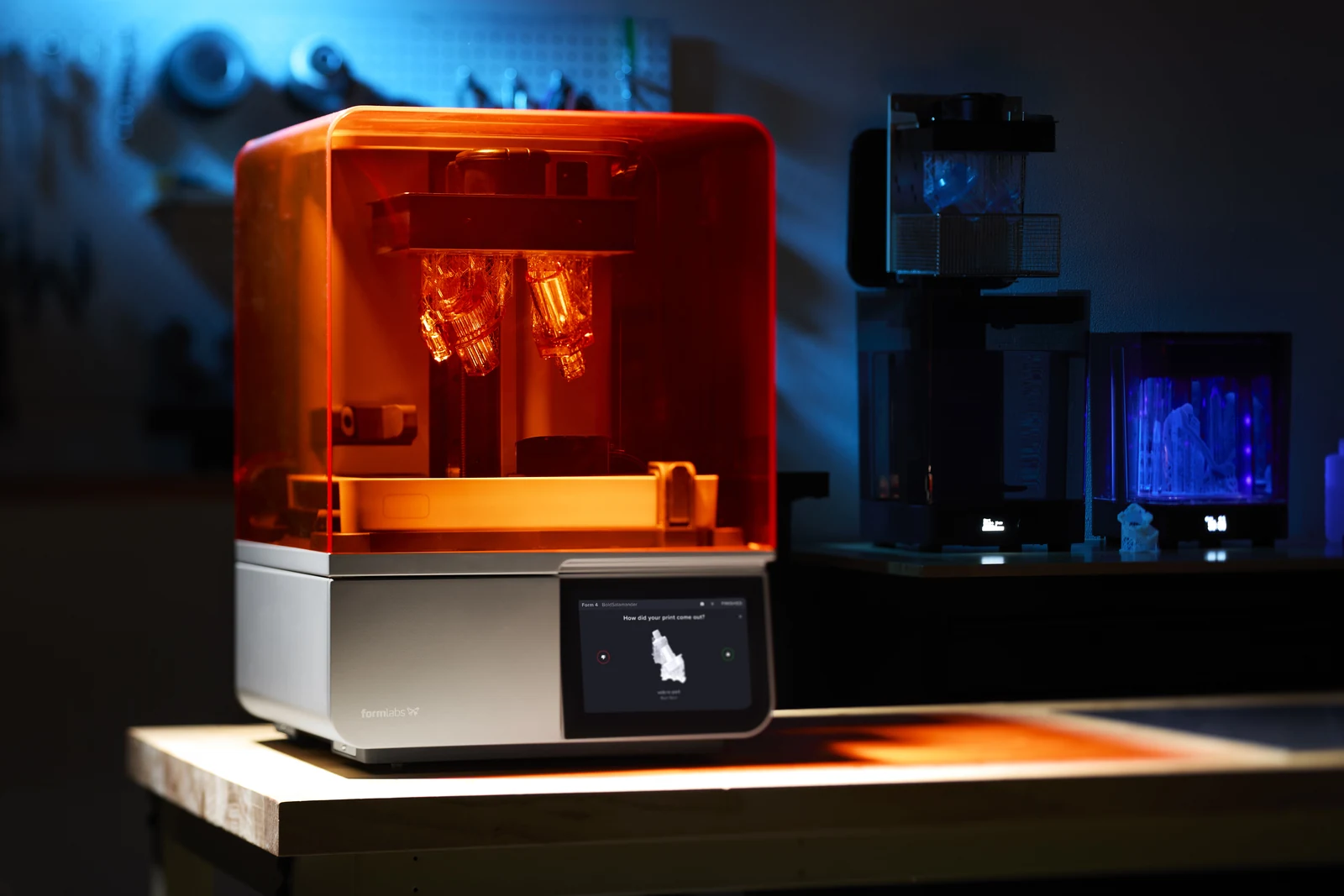 Formlabs Form 4 resin 3D printer shows parts on its build platform under an orange cover in a dim workshop