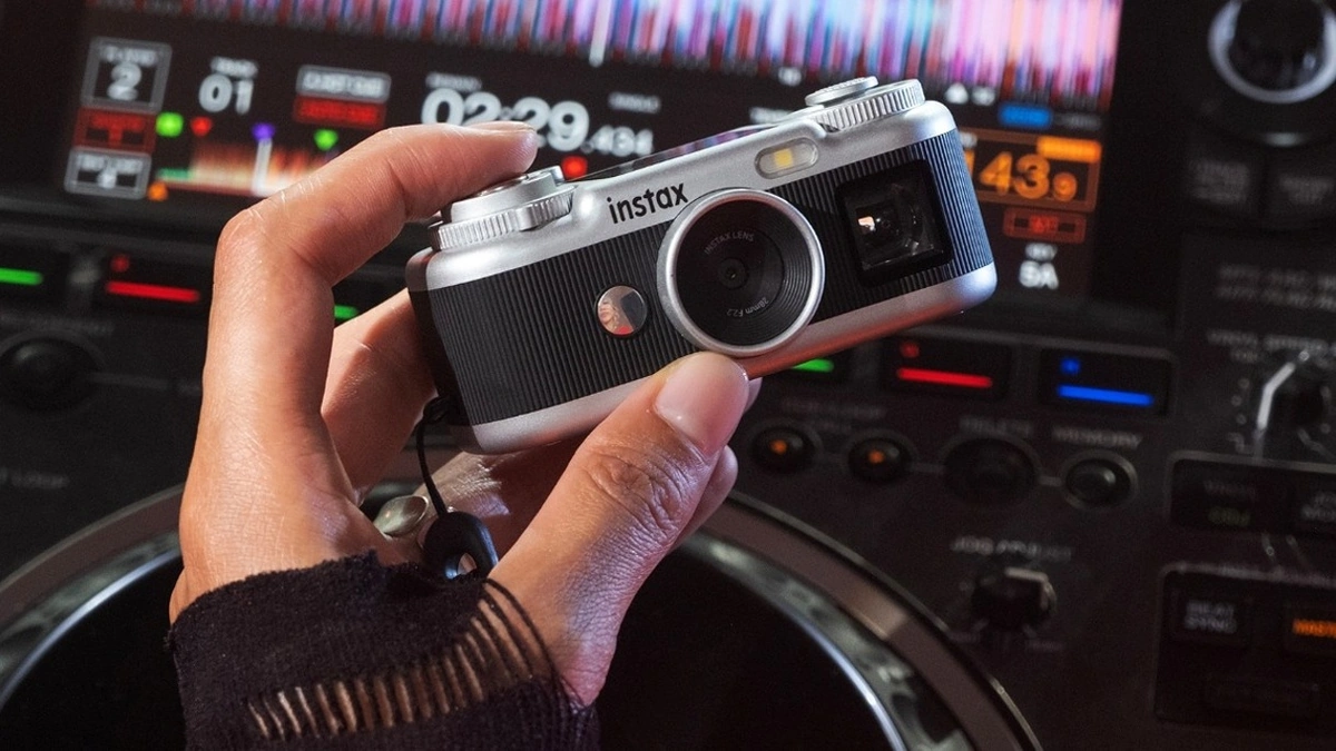 Hand holding a silver Instax Pal 2 camera in front of illuminated DJ equipment