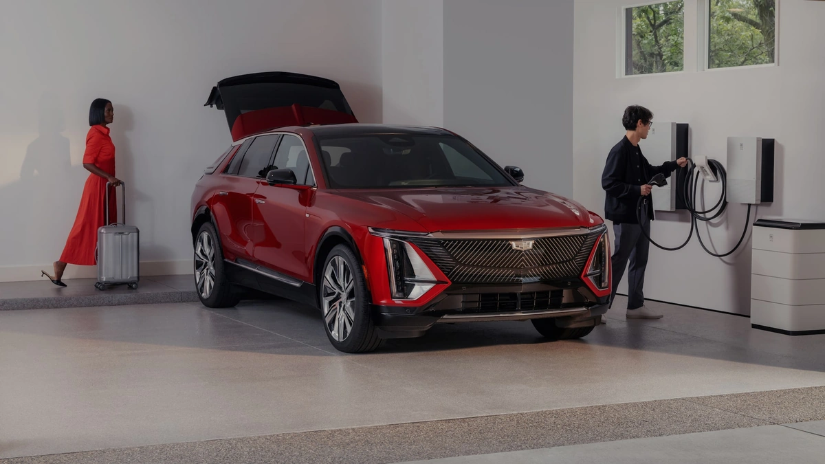 Red Cadillac electric SUV in a garage beside a wall charger and home energy units with two people nearby