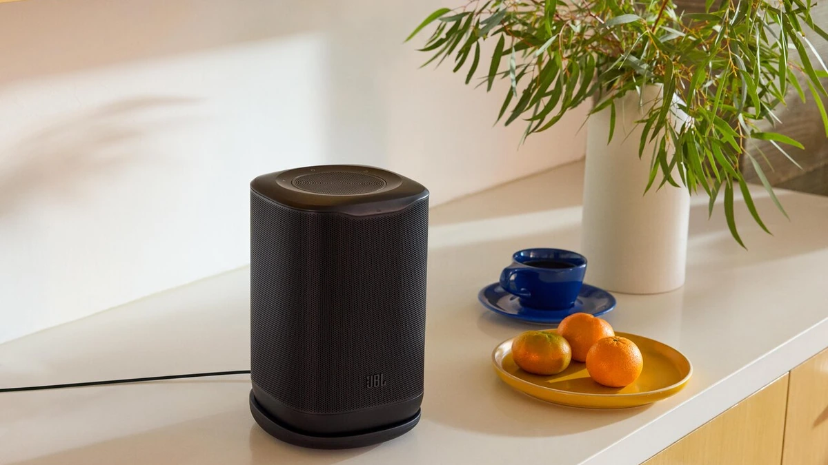Black JBL Cove P1 speaker on a white sideboard beside a blue cup, a plate of oranges and a vase of eucalyptus