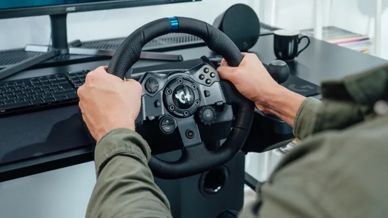 Logitech G racing wheel held by a player's hands at a desk with a monitor and keyboard behind it