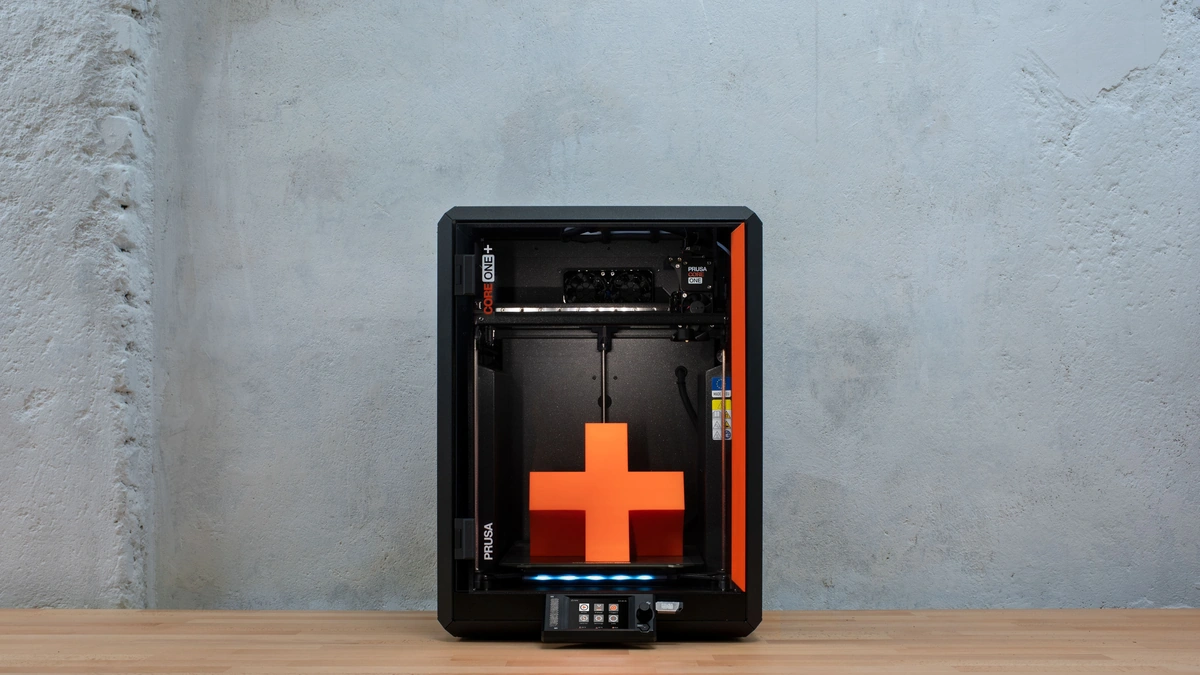 Prusa CORE One+ enclosed 3D printer with an orange cross print inside on a wooden table against a concrete wall