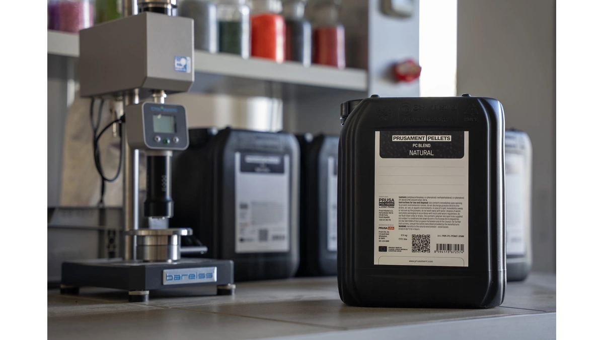Black Prusament Pellets PC Blend canister on a lab bench beside a hardness tester and more canisters