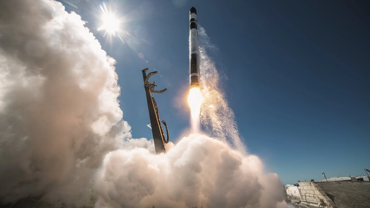 Rocket Lab Electron rocket lifting off with a plume of smoke and flame against a blue sky