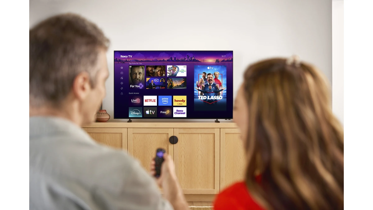Roku Pro Series OLED TV on a wood console showing the Roku home screen as two people watch