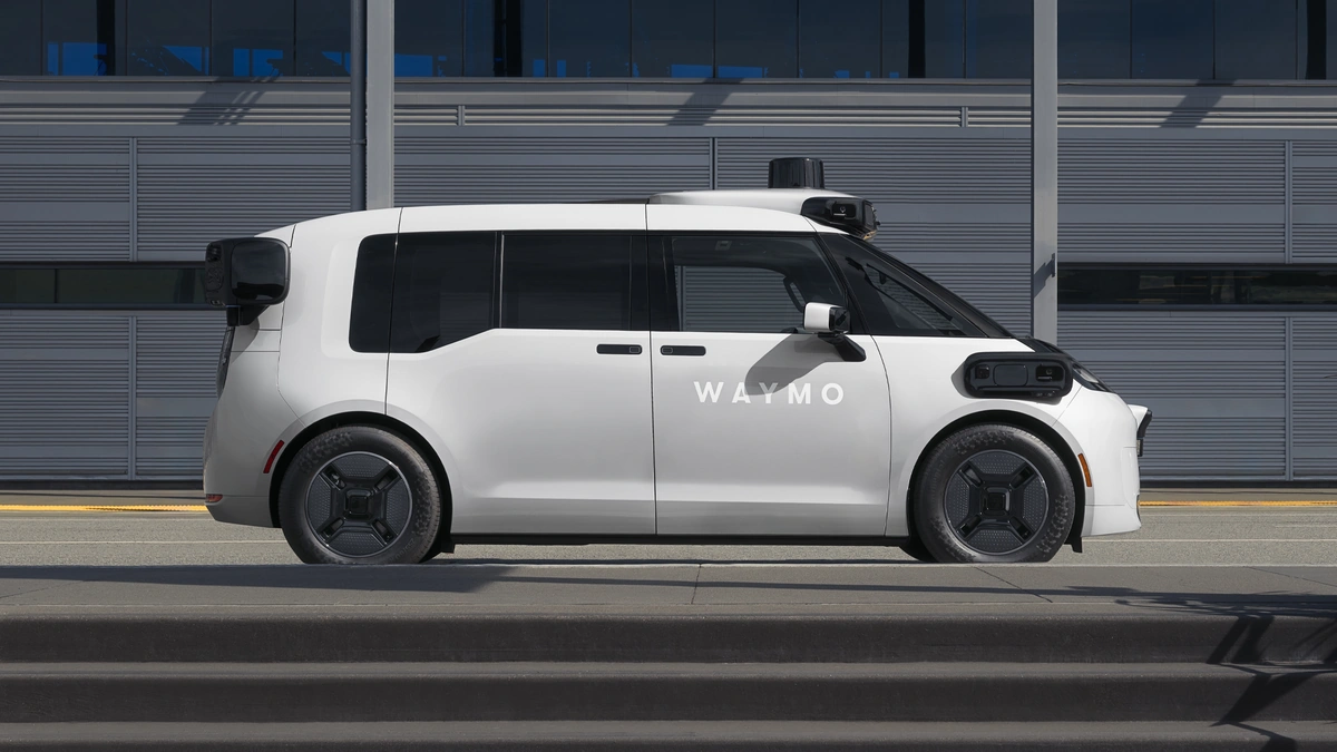 White Waymo robotaxi with roof-mounted sensors, seen side-on in front of a gray paneled wall